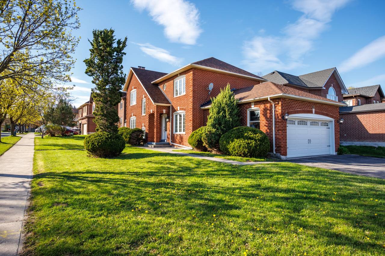 83 John Stiver Crescent seen from the sidewalk on a clear spring morning, a two-storey red brick house with a white front door, an attached garage to the right, and a wide lawn running down to the street.